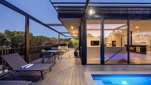 Een modern terras buiten met lounge fauteuils, een zwembad en een overdekte eethoek met glas.