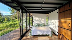 Een slaapkamer met een groot bed, een houten vloer en uitzicht op een bos door glazen deuren.