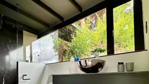 Un baño moderno con suelo de madera, inodoro, lavamanos y un ventanal con vistas a zonas verdes.