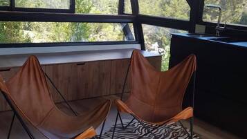 Una habitación con un alfombra estampado con motivos de cebra, dos sillas marrones, un armario de madera y vistas a la vegetación a través de grandes ventanales.