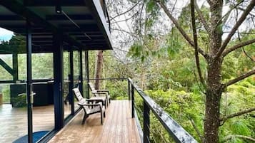 Une terrasse en bois avec des chaises, entourée d’arbres et dotée d’une rambarde en verre.