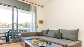 A living room with a sofa, a wooden table with food, a sliding glass door, and a view of greenery outside. - Apartment Odisea by Interhome (Lloret de Mar)