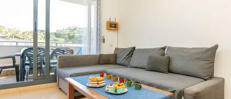 A living room with a sofa, a wooden table with food, a sliding glass door, and a view of greenery outside.