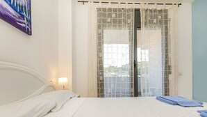 A bedroom with a bed, a nightstand, a lamp, and a view through a window with a patterned curtain. - Apartment Odisea by Interhome (Lloret de Mar)