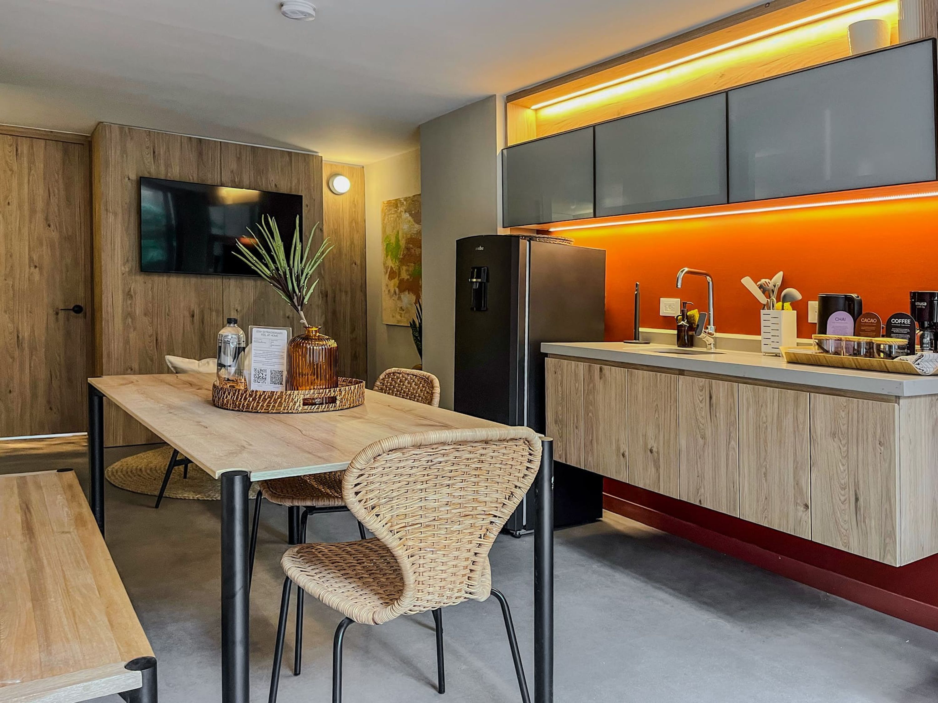 Una cucina moderna con un tavolo da pranzo in legno, sedie in vimini, TV a incasso e frigorifero.