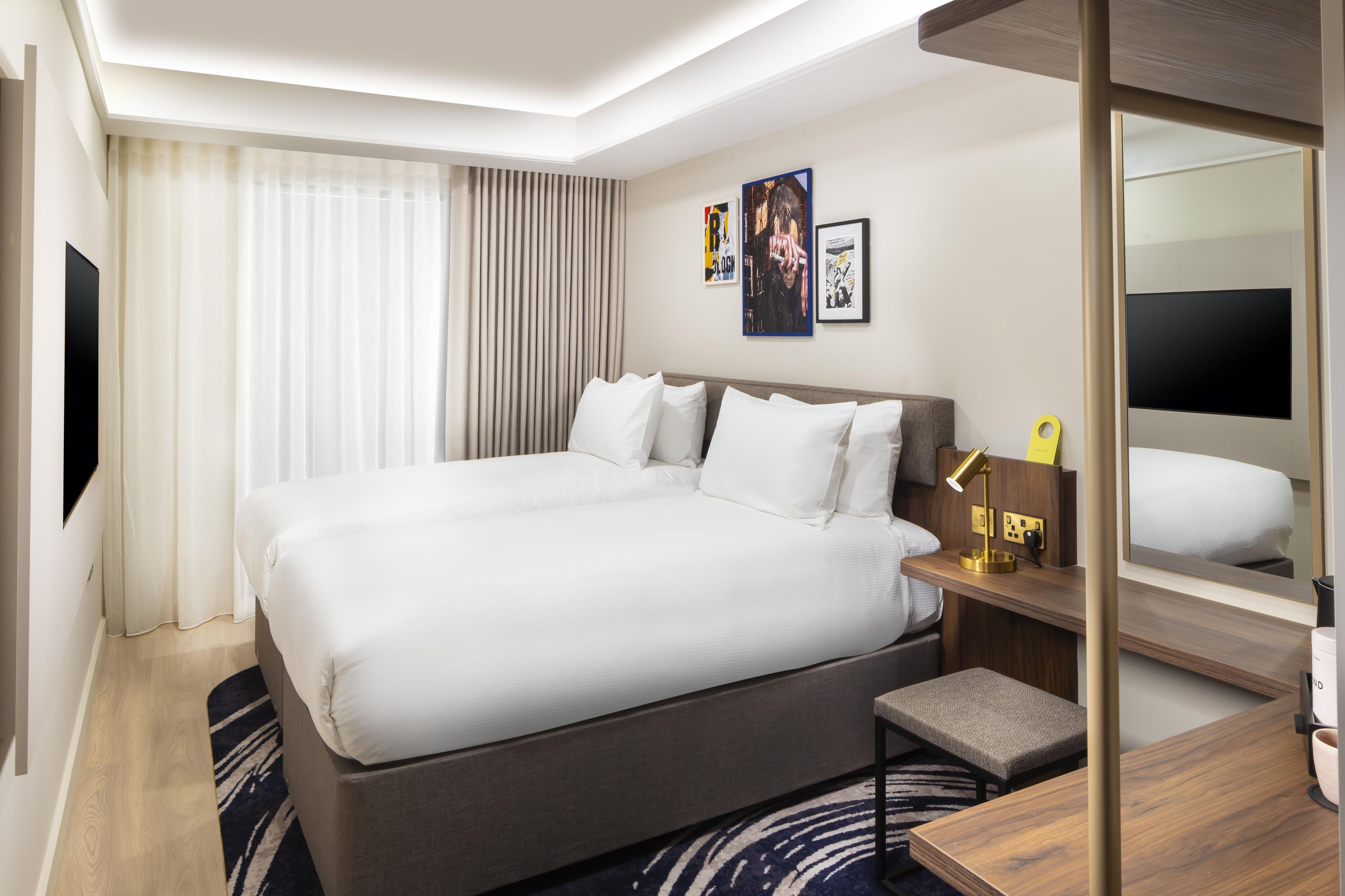 Ein Hotelzimmer mit einem großen Bett, einem Nachttisch mit Lampe, einem kleinen Hocker, einem Spiegel und Bildern an der Wand.