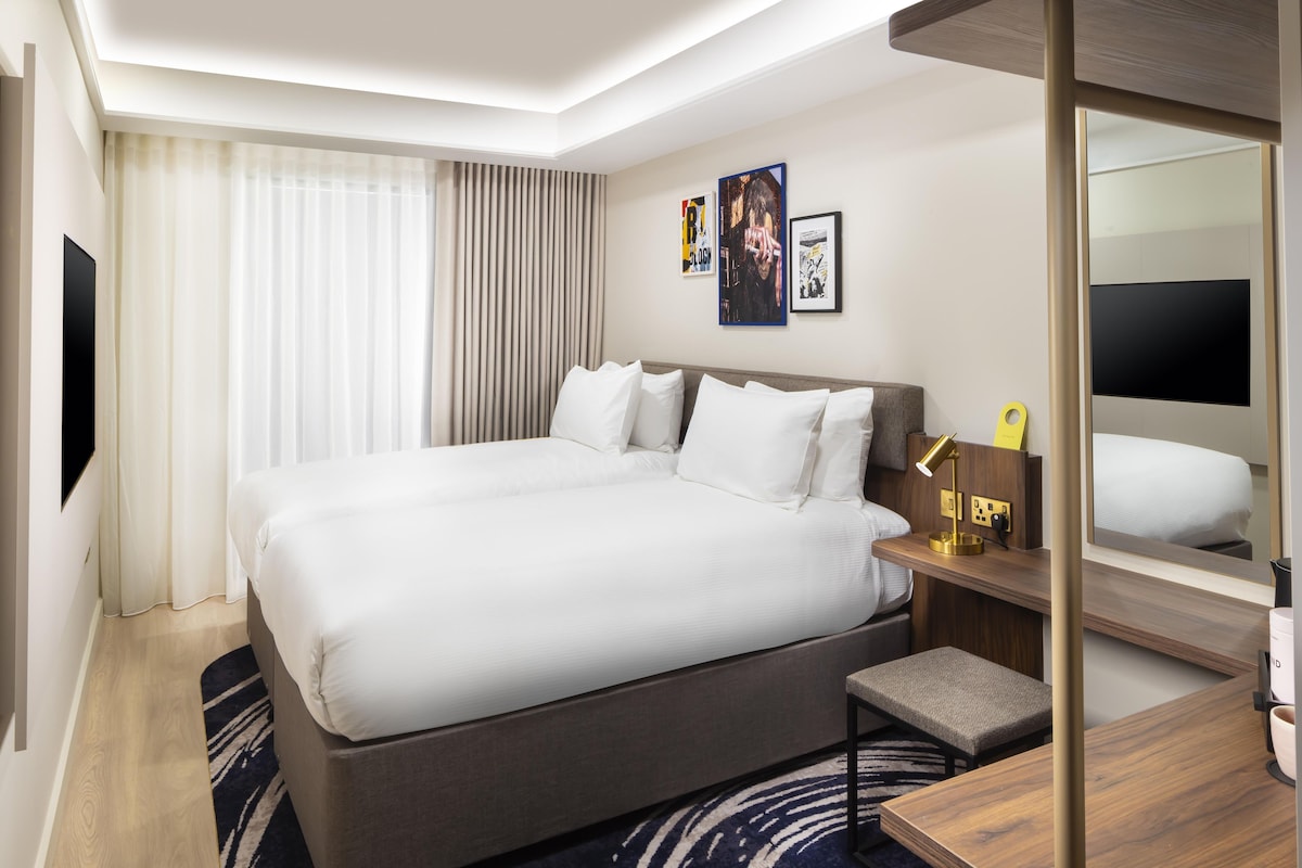 A hotel room with a large bed, a bedside table with a lamp, a small stool, a mirror, and artwork on the wall.