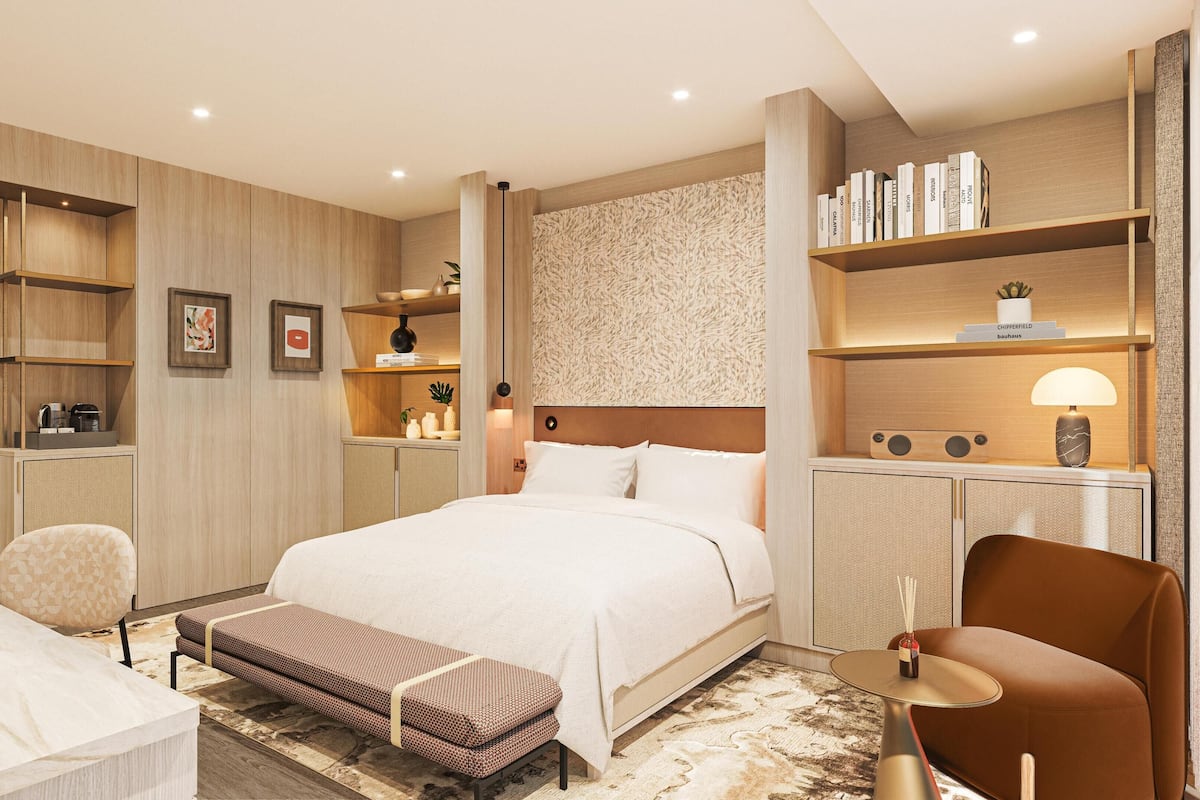 A modern hotel room with a large bed, a sitting area with a bench, a desk, and a bookshelf.