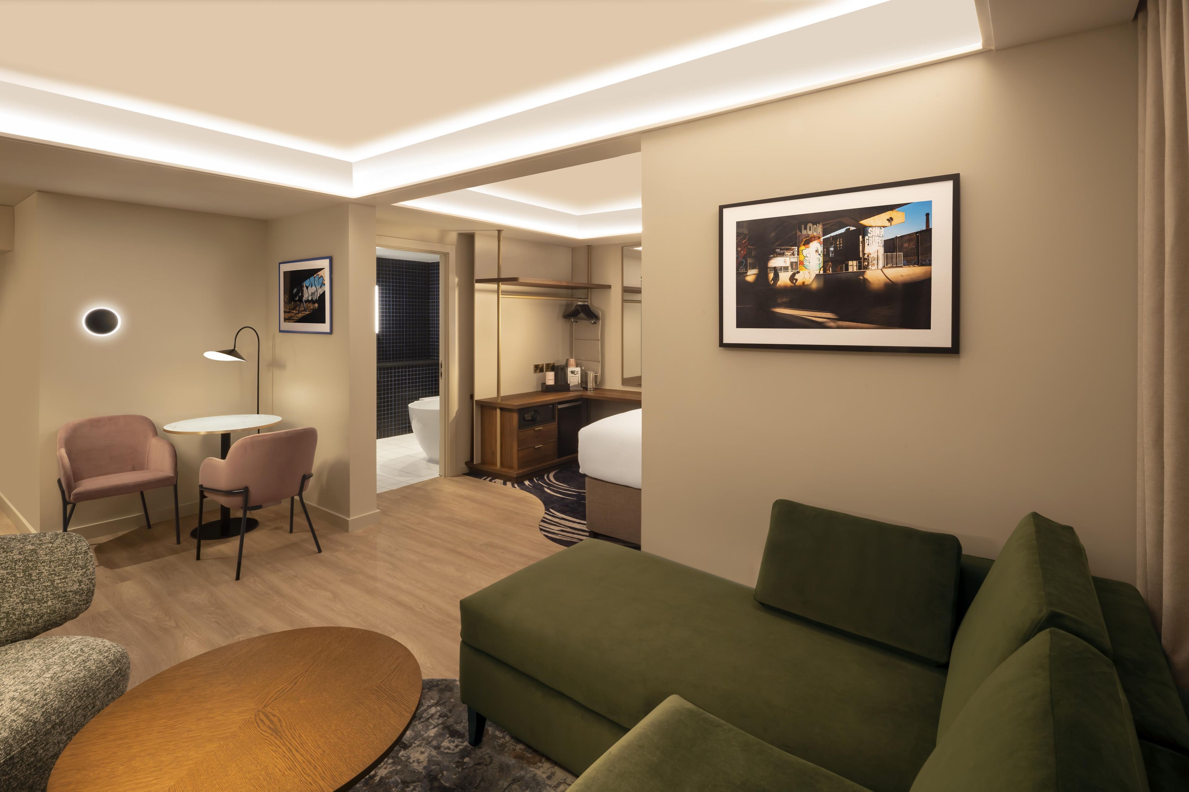 Una habitación de hotel moderna con un sofá verde, una pequeña mesa redonda, un escritorio con una silla, un espejo circular y un cuadro enmarcado en la pared.