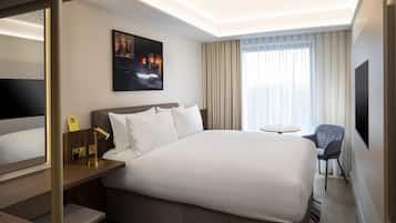 A hotel room with a large bed, a desk with a lamp, a chair, a small table, a TV, and a window with curtains.
