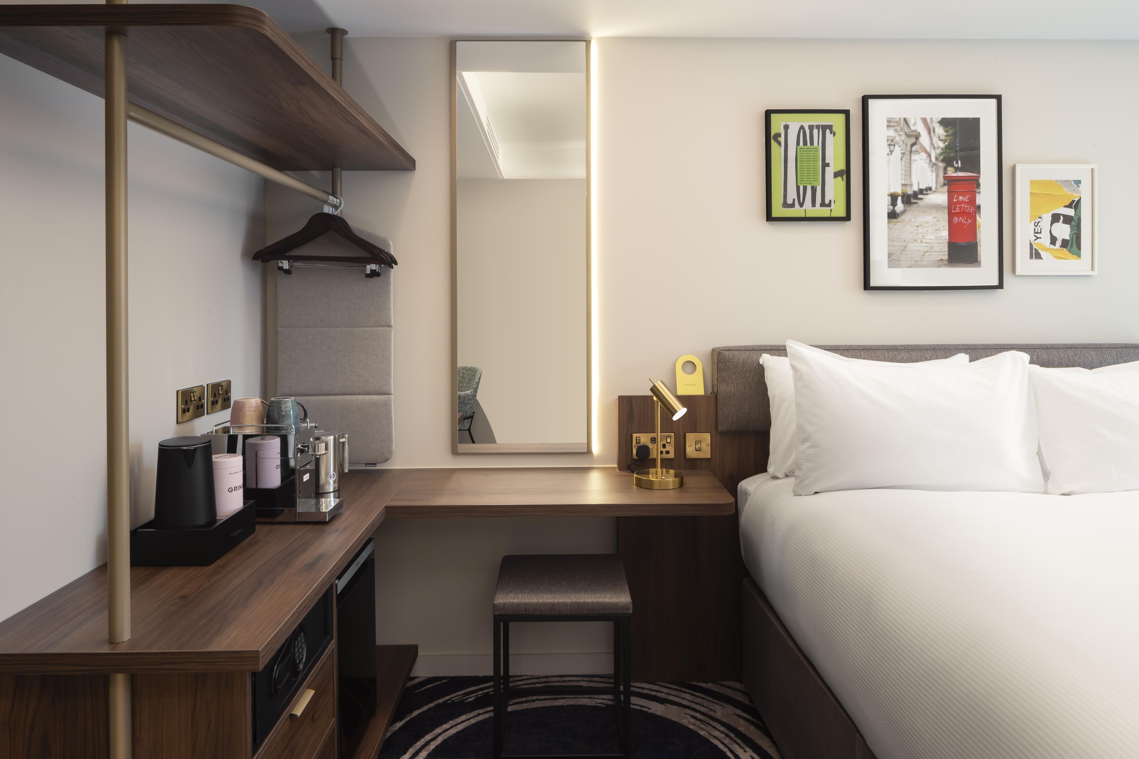 Een hotelkamer met een bed, een bureau met een koffieapparaat, een kapstok, een klein krukje en een spiegel.