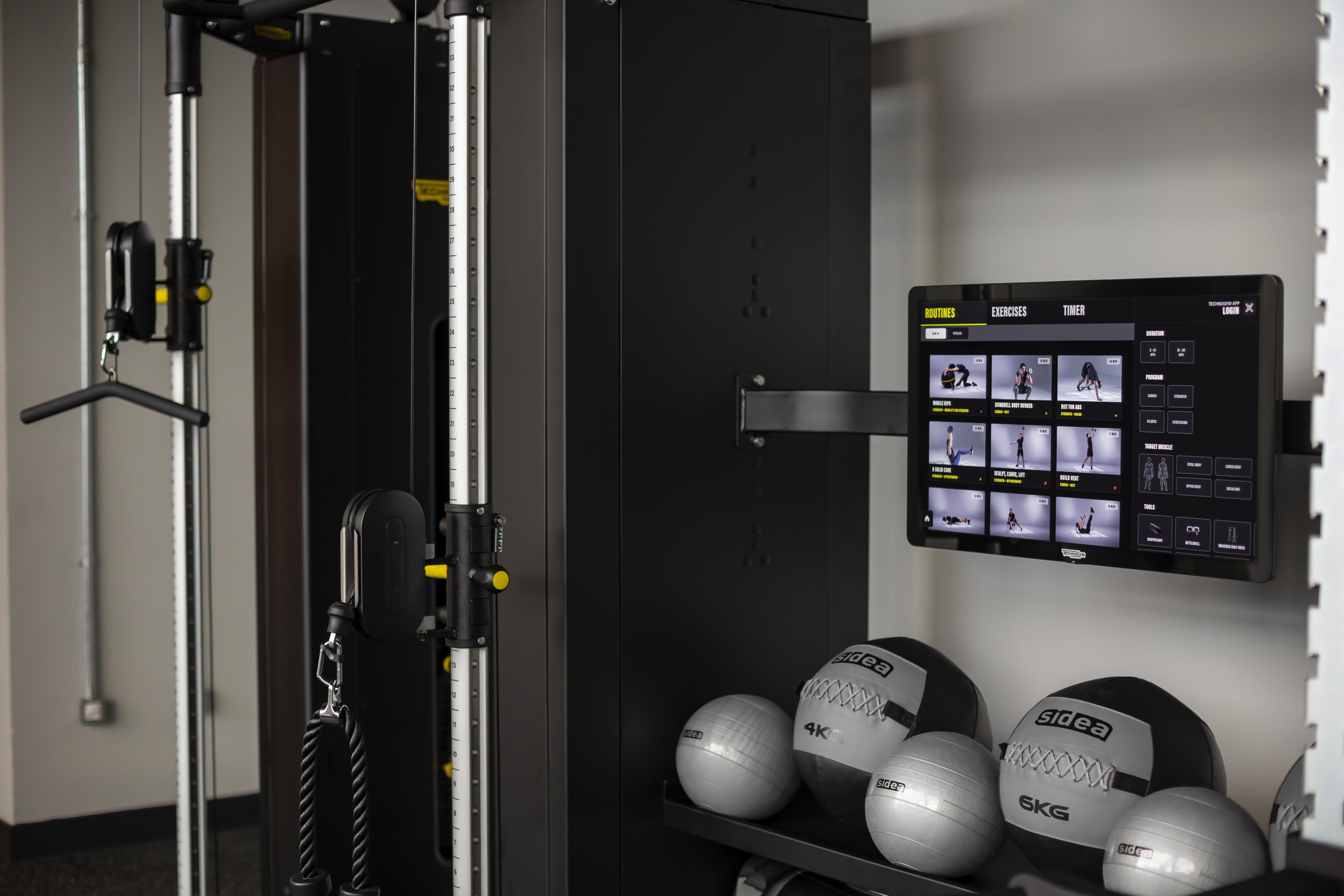 A gym with exercise equipment, a digital screen displaying workout routines, and weightlifting balls.