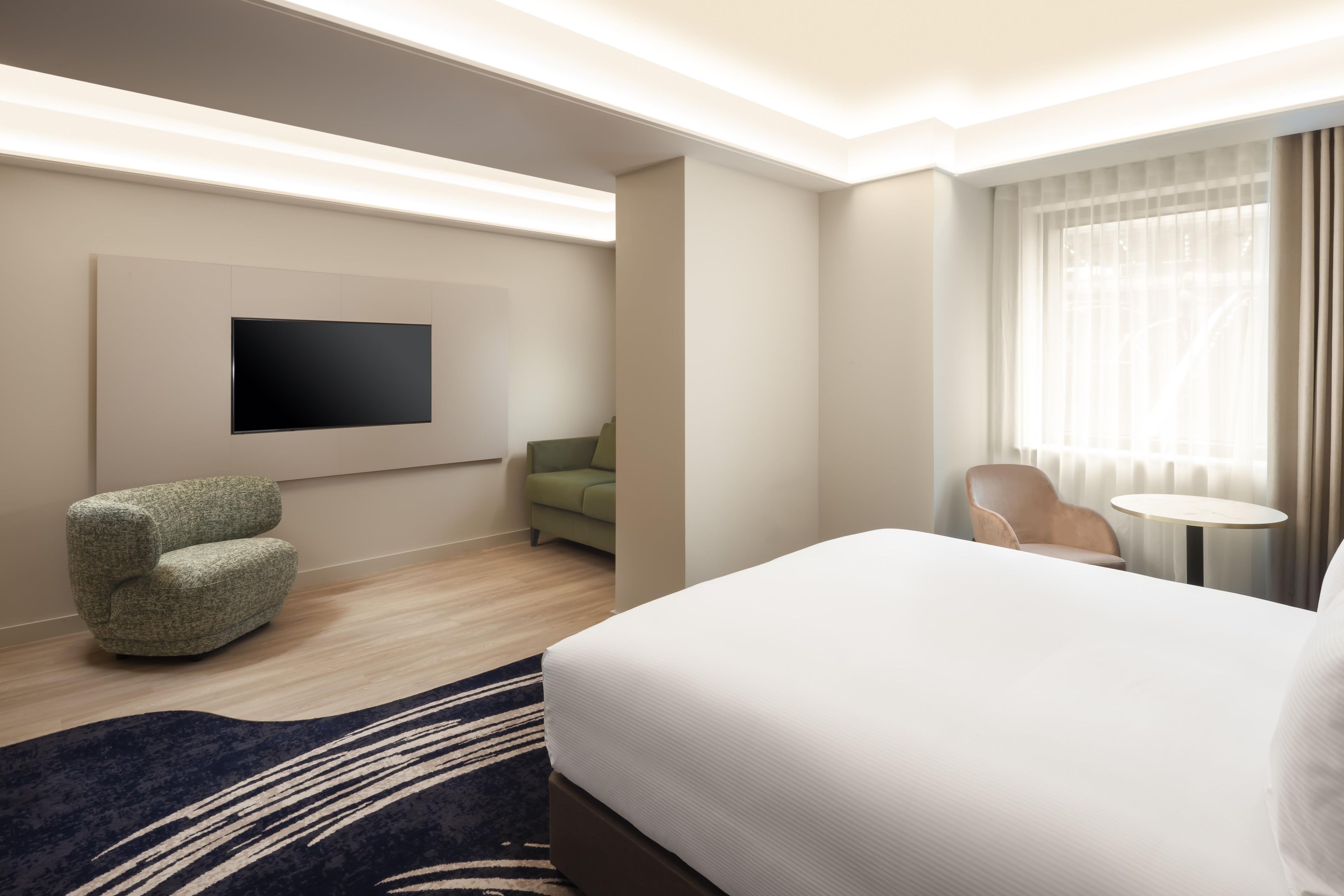 Een hotelkamer met een bed, een flatscreen televisie, een groene bank, een stoel, een klein rond tafeltje en een raam met gordijnen.