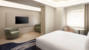 A hotel room with a bed, a flat-screen TV, a green sofa, a chair, a small round table, and a window with curtains.