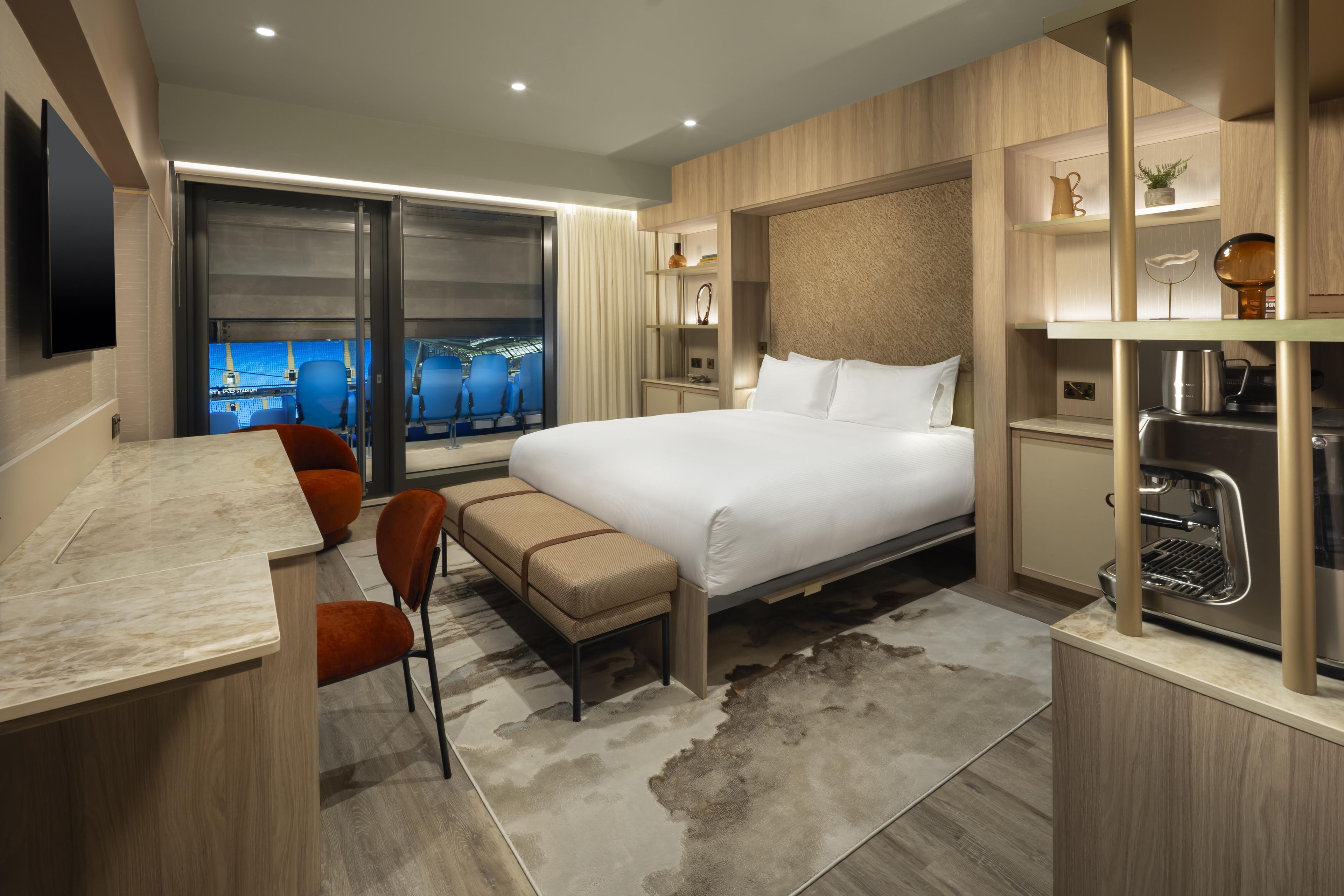 A modern hotel room with a bed, a desk, a chair, a sofa, and a view of a stadium.