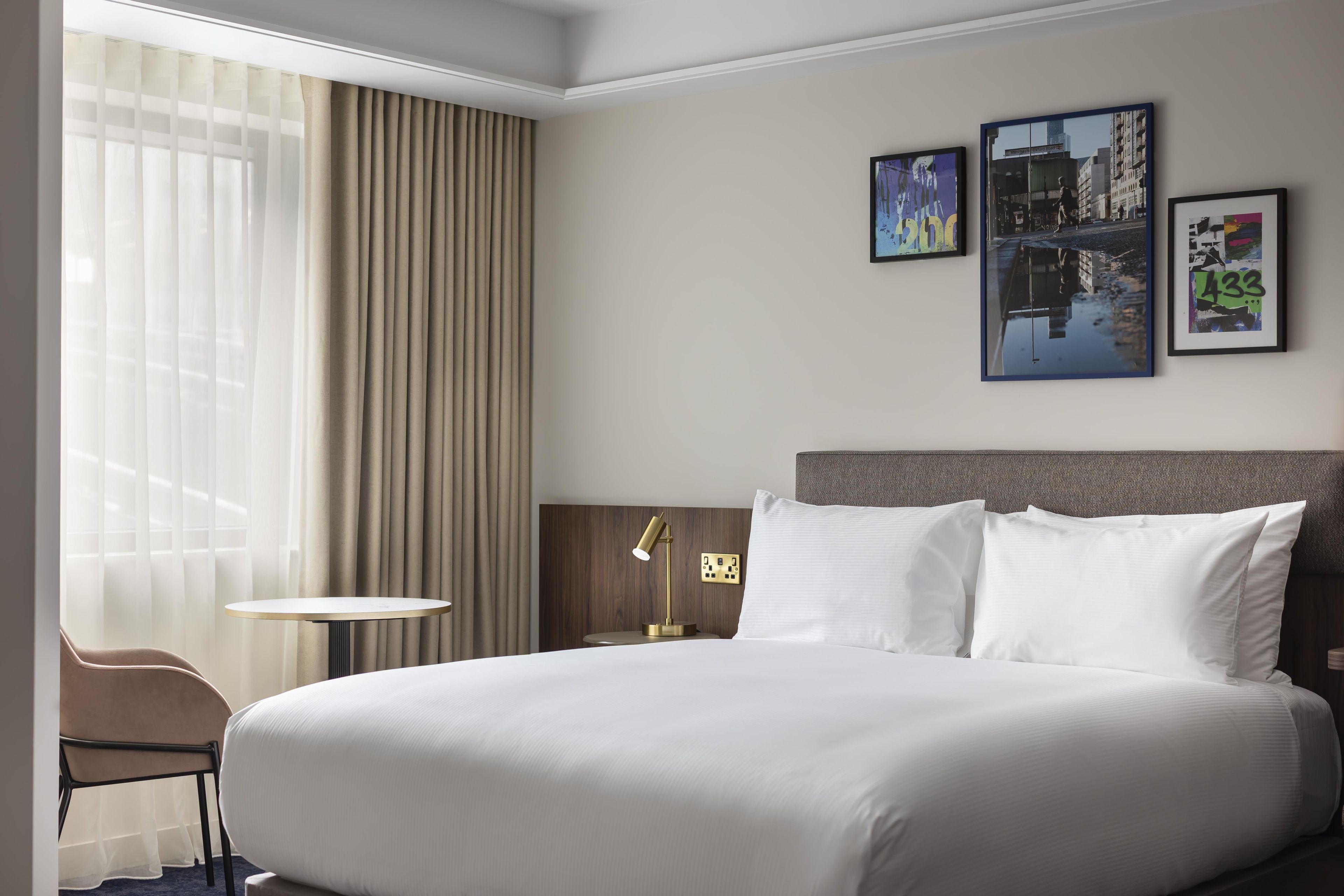 Camera d'albergo con un letto grande, un tavolino e due sedie. In parete, tre quadri incorniciati.