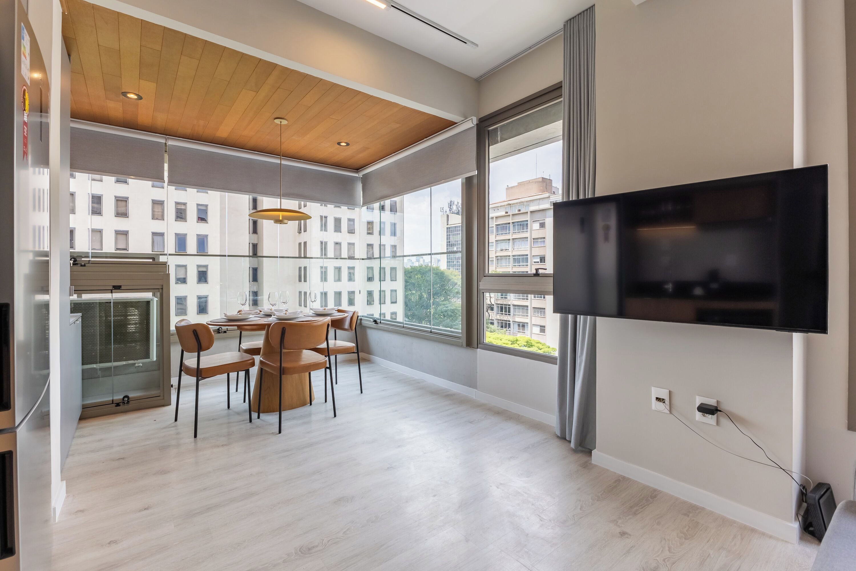 Un vaste espace de vie comprenant une table à manger, des chaises, un téléviseur fixé au mur et de grandes fenêtres offrant une vue sur le paysage urbain.