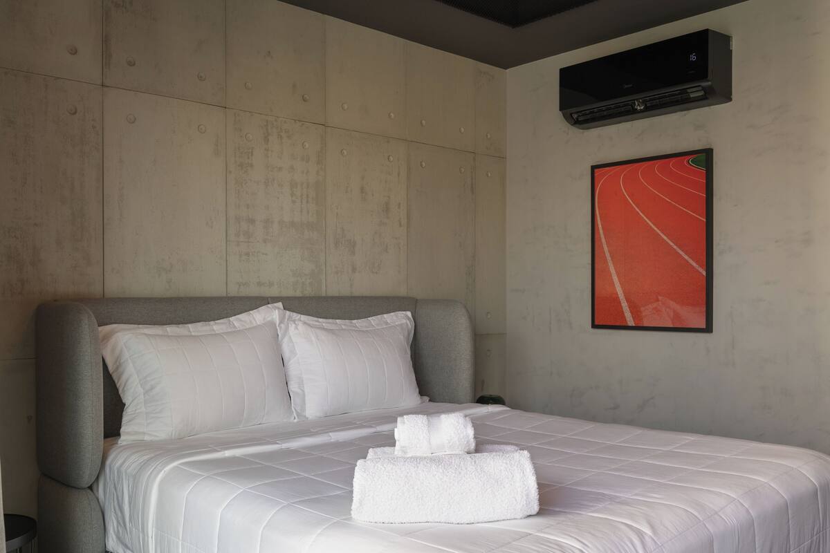 A hotel room with a bed, pillows, a wall-mounted air conditioner, and a framed picture.