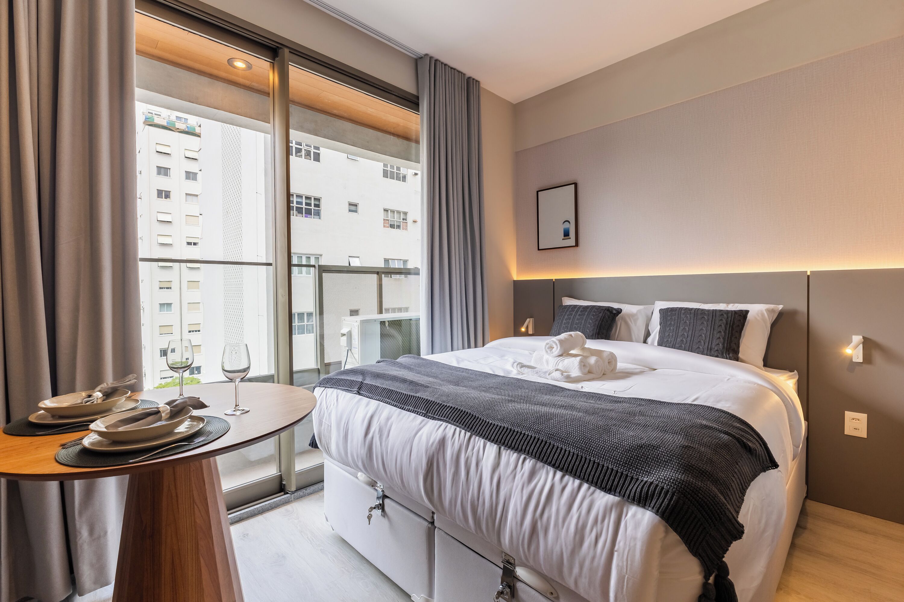 A hotel room with a bed, a dining table with plates and glasses, a window with curtains, and a city view.