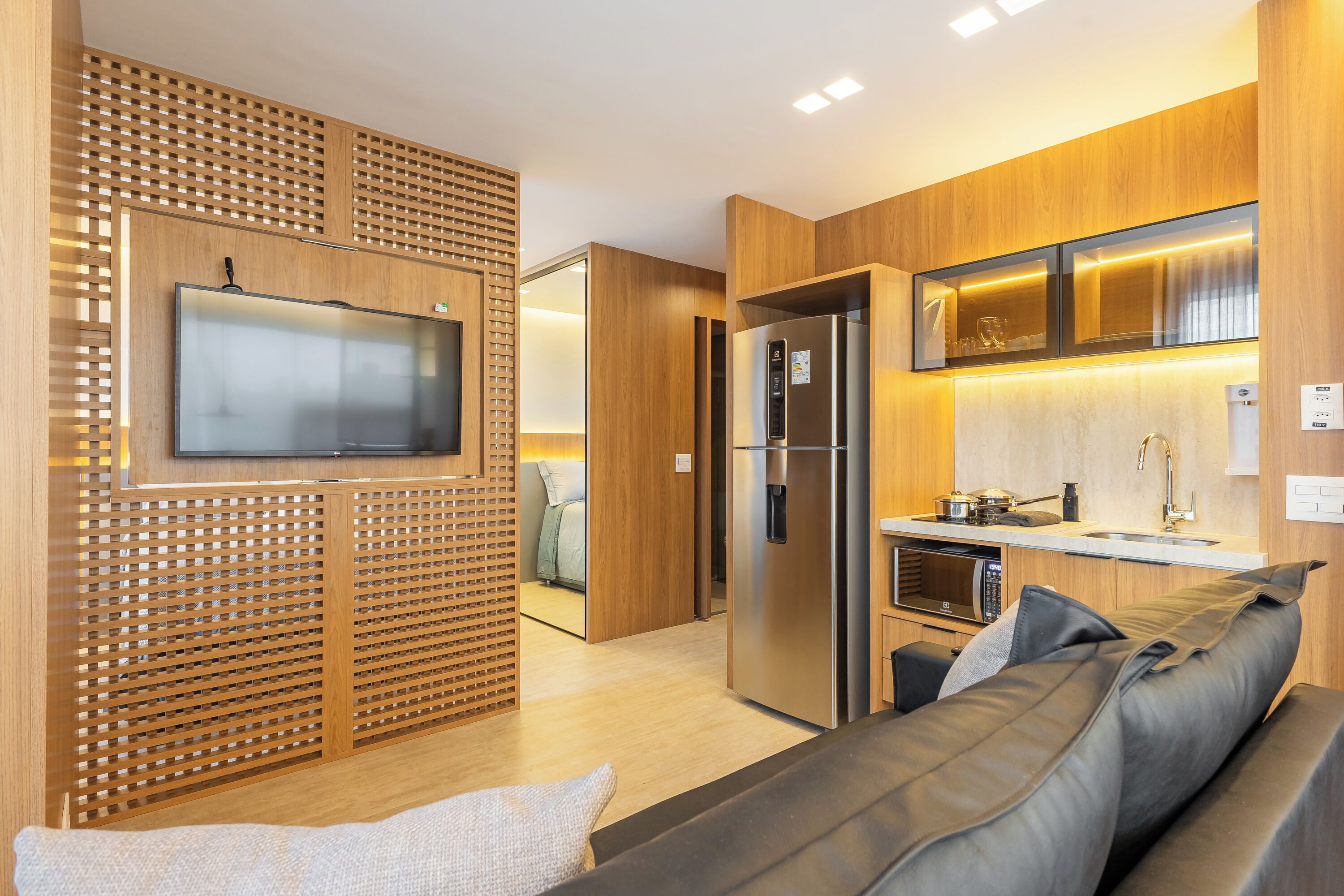 Cocina moderna con refrigerador, microondas y estufa integrados, muebles de madera y un televisor de pantalla plana montado en un panel de madera.