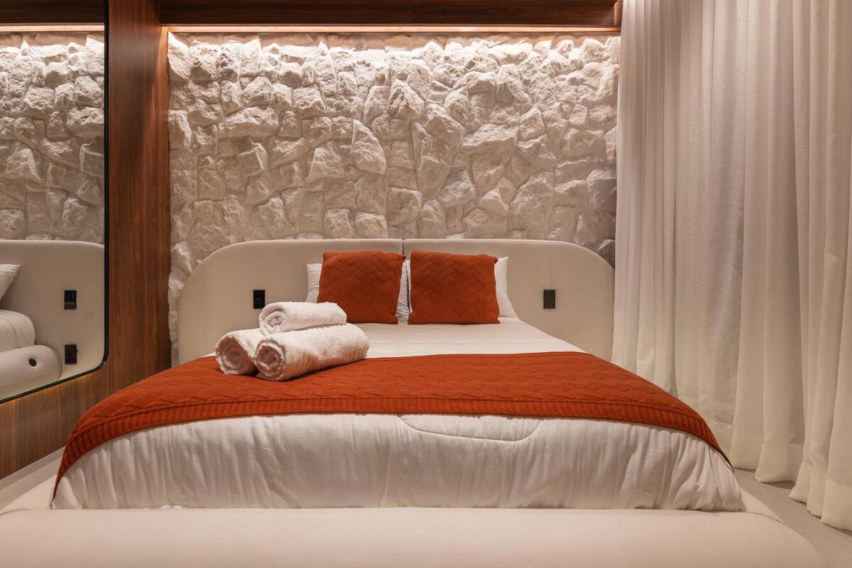 A bed with white bedding and orange pillows, a stone wall behind, and a white curtain to the right.
