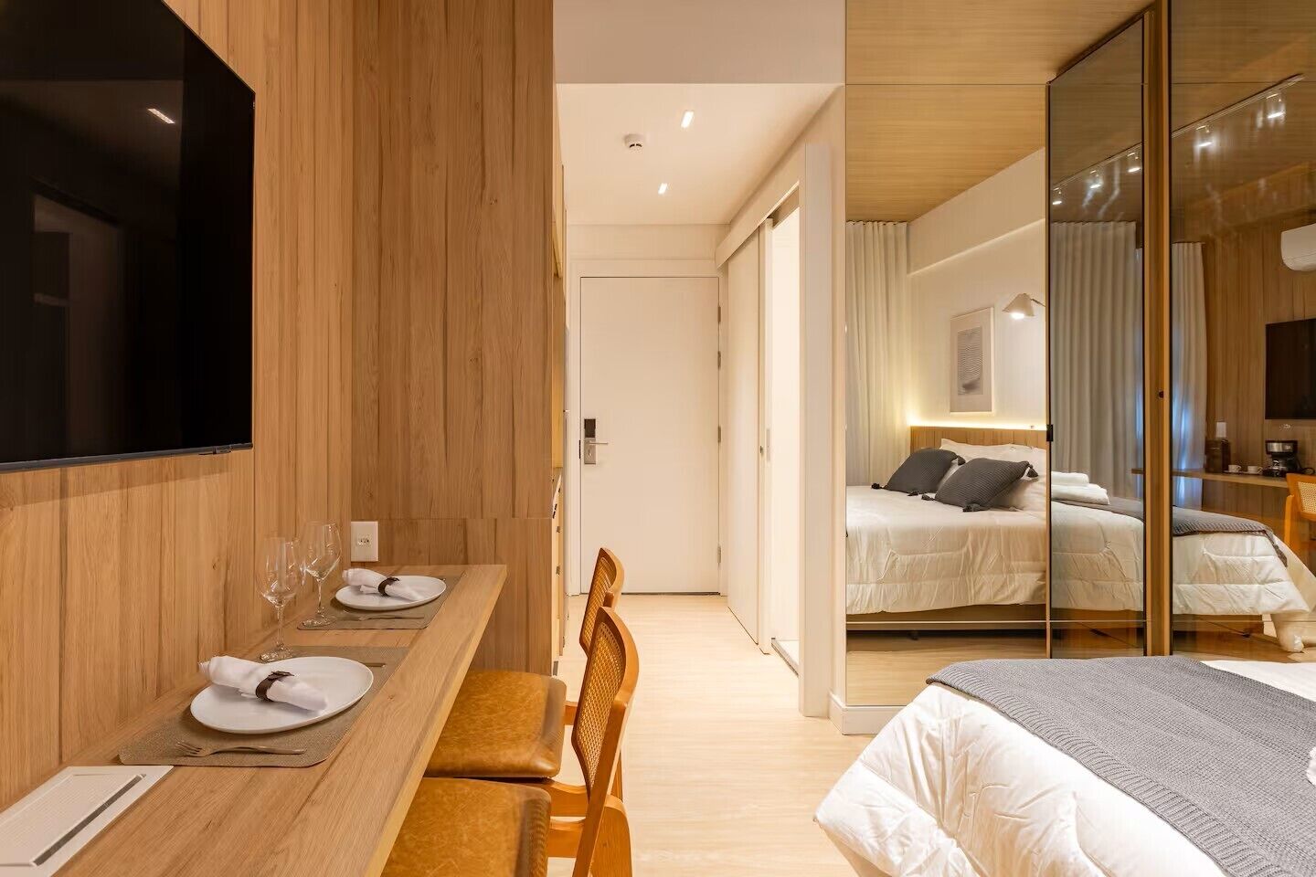 Una habitación de hotel moderna con una zona de comedor de madera, una cama con cabecero y un espejo grande.