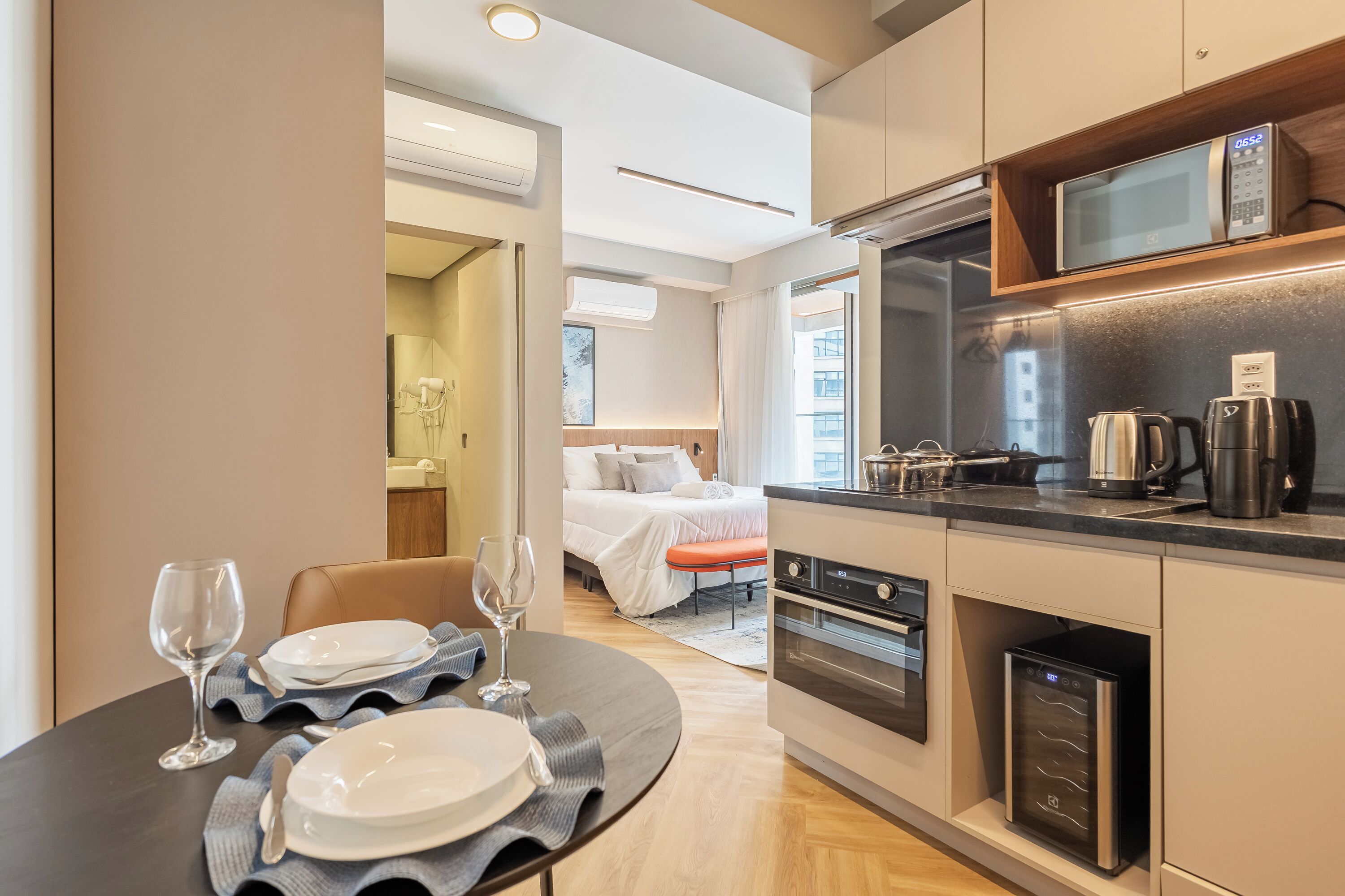 A modern hotel room with a kitchen area, a dining table set with plates and glasses, and a bed with a white comforter.