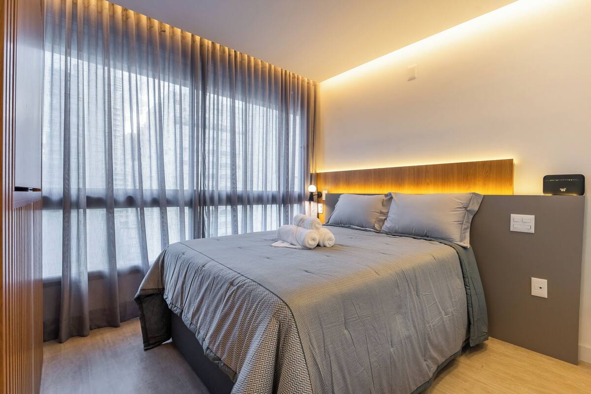 Une chambre d’hôtel moderne dotée d’un grand lit, d’une tête de lit avec éclairage intégré, d’une table de chevet et d’une fenêtre ornée de rideaux légers.