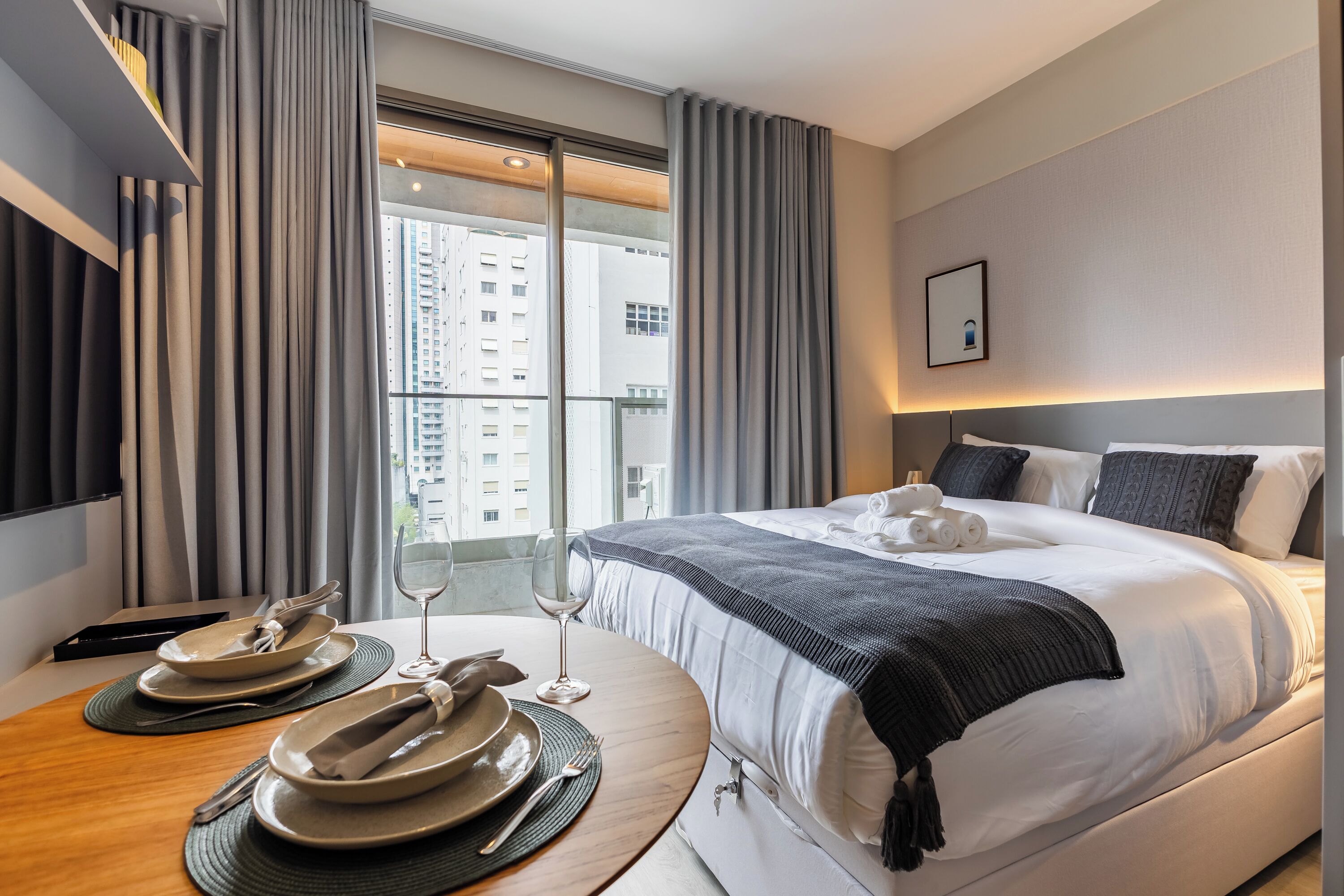 Habitación de hotel con una cama bien hecha, una mesa de comedor con platos y cubiertos, una copa de vino y vistas a la ciudad a través de amplios ventanales.