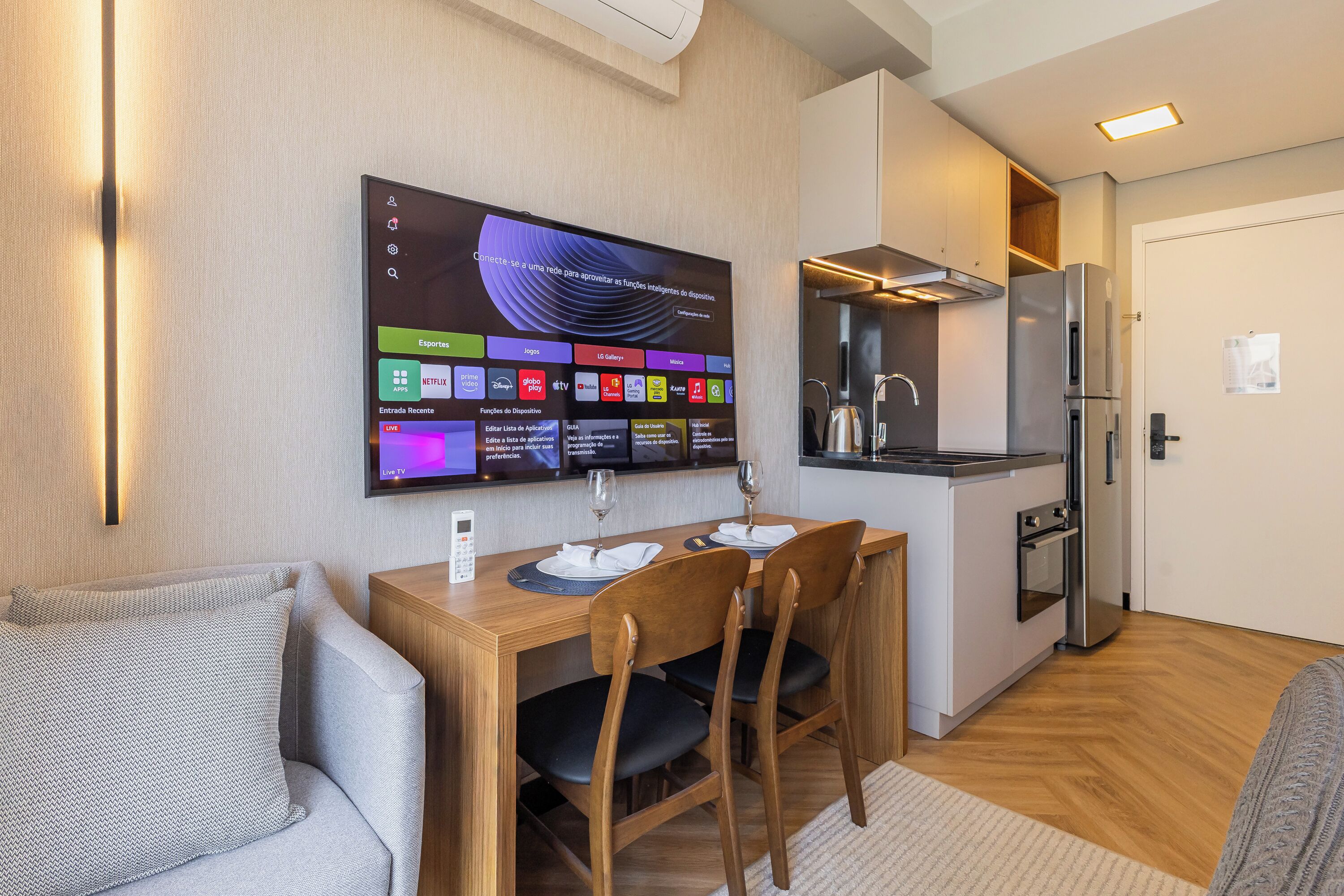 Eine moderne Küche mit Einbauofen und Kühlschrank, ein Essbereich mit Holztisch und -stühlen sowie ein an der Wand befestigter Fernseher, der verschiedene Streaming-Dienste anzeigt.