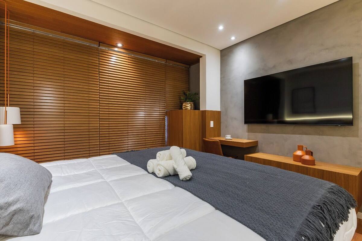 A modern hotel room with a large bed, a flat-screen TV mounted on the wall, a wooden headboard, and a small desk with a chair.