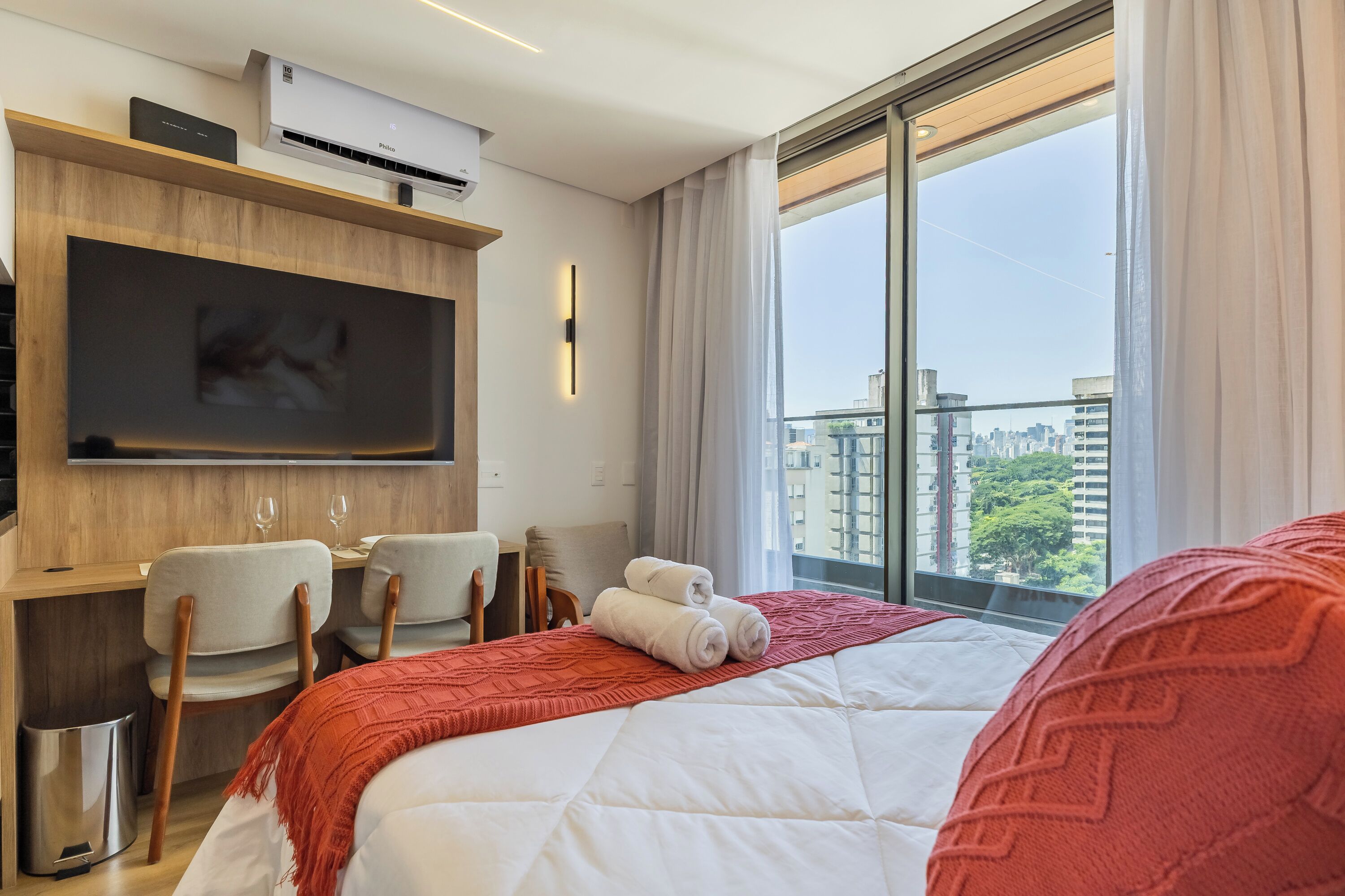 A hotel room with a bed, a TV mounted on the wall, a desk with chairs, and a large window with a city view.