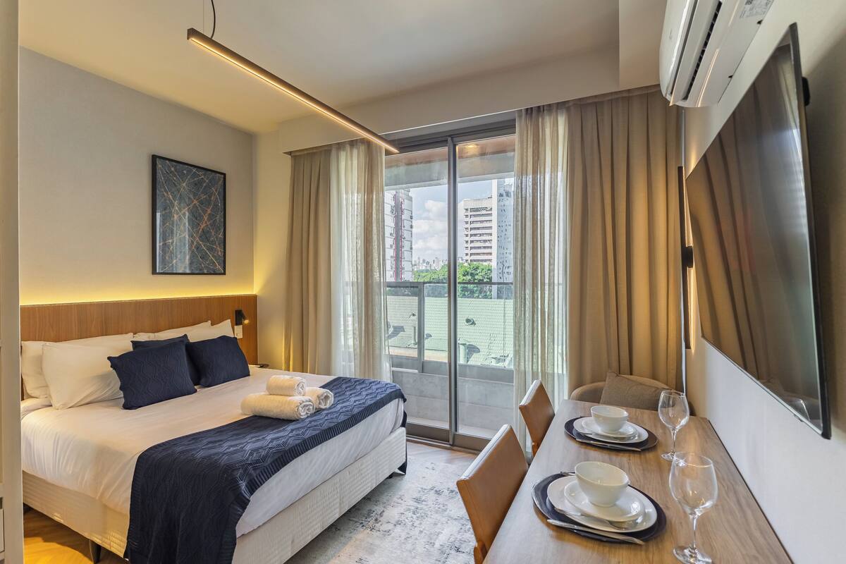 A hotel room with a bed, a dining table with chairs, a large window with curtains, and a mounted TV.