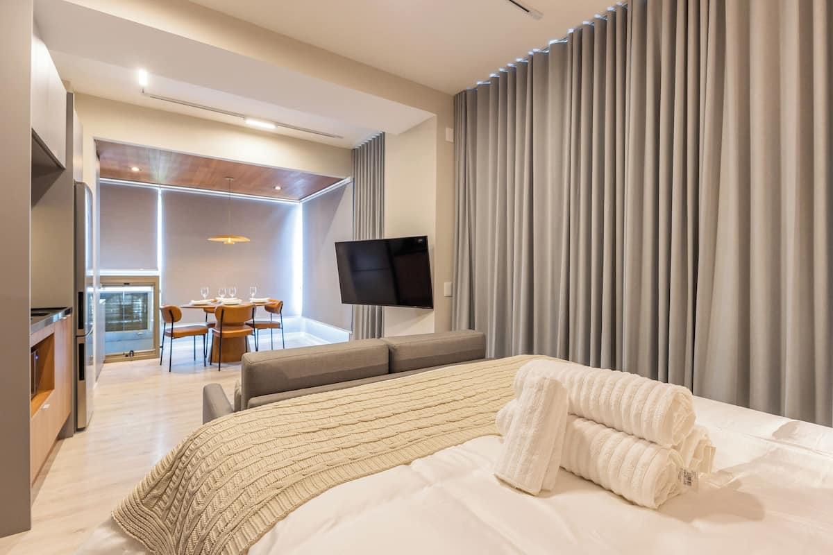 Une chambre d’hôtel moderne dotée d’un grand lit, d’un téléviseur à écran plat, d’un coin repas avec une table et des chaises, et offrant une vue sur la ville grâce à de grandes fenêtres.