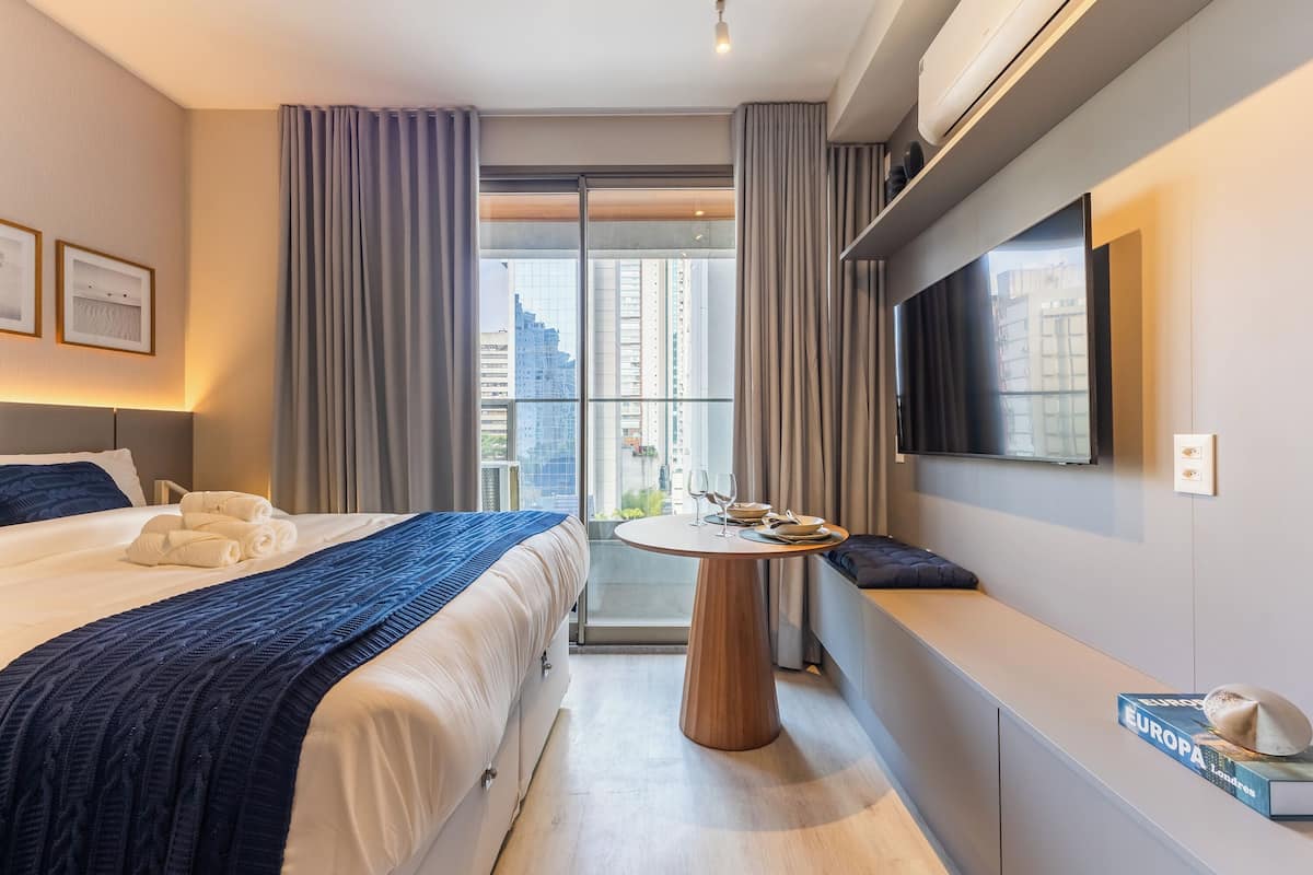 Une chambre d’hôtel moderne avec un grand lit, une table ronde sur laquelle sont posés un livre et un chapeau, une télévision et un balcon avec des rideaux.