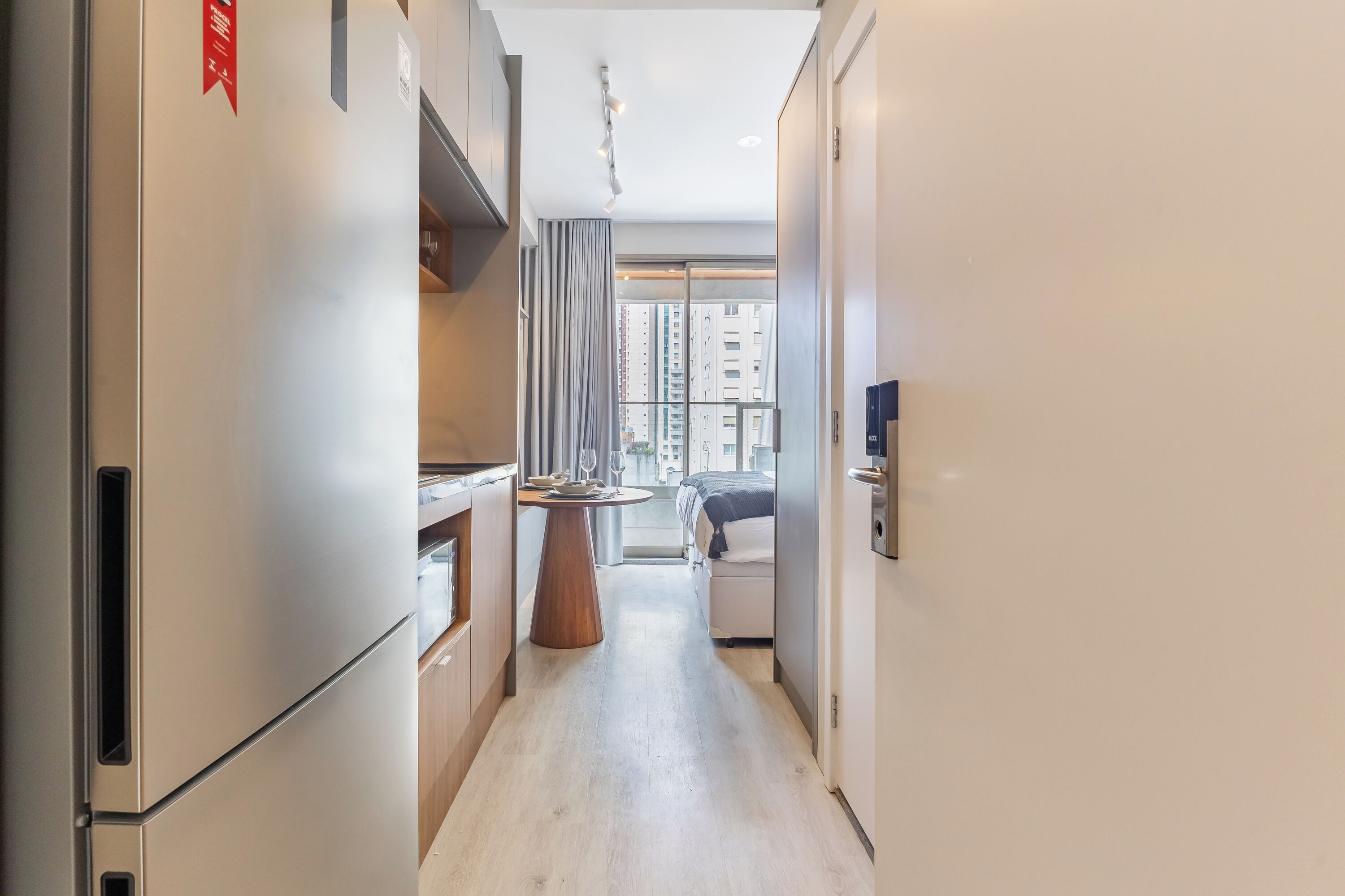 A narrow hallway with a kitchen area on the left, a dining table in the center, and a bed with a view to the outside on the right.