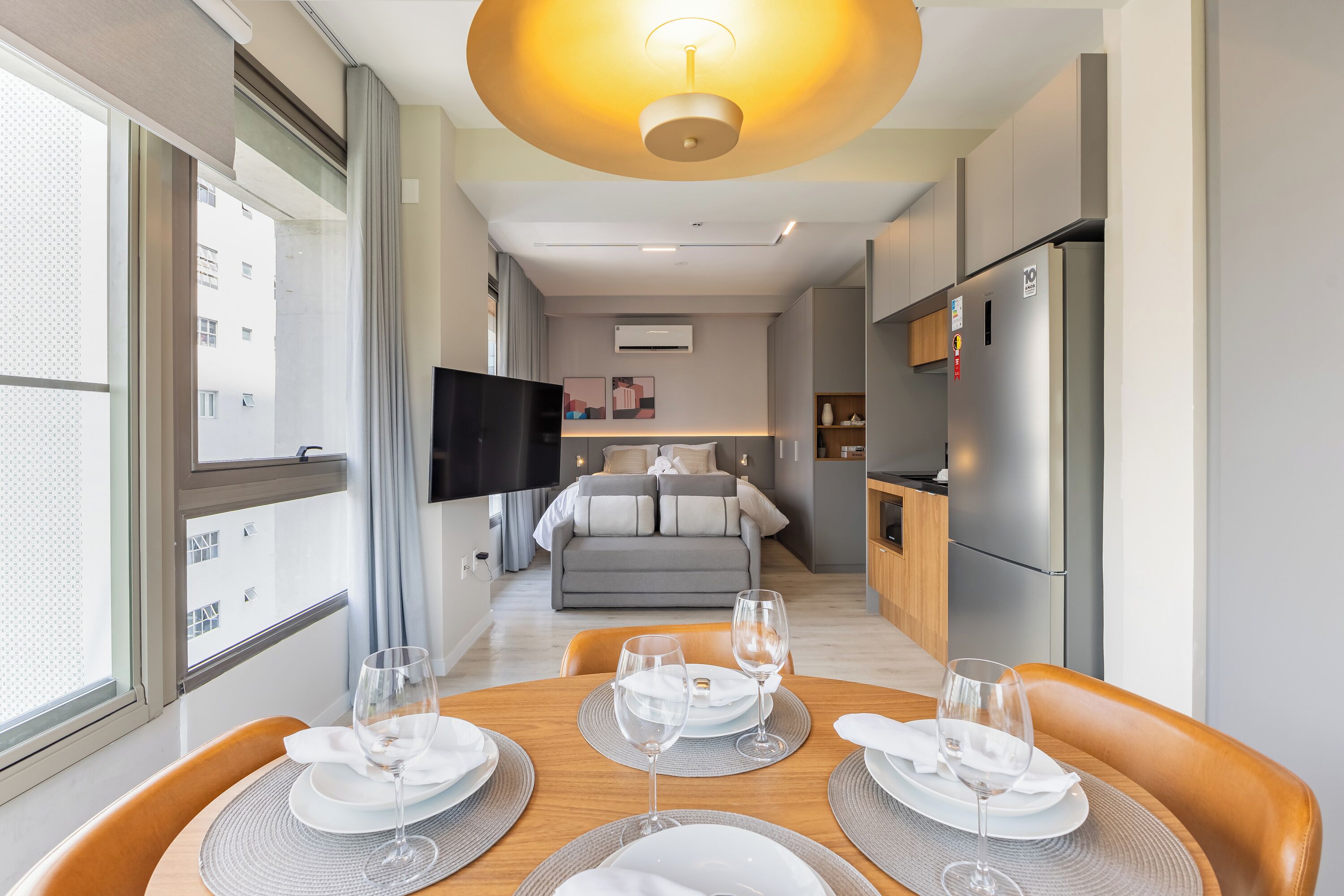 Una habitación de hotel moderna con zona de comedor, kitchenette y un televisor al fondo.