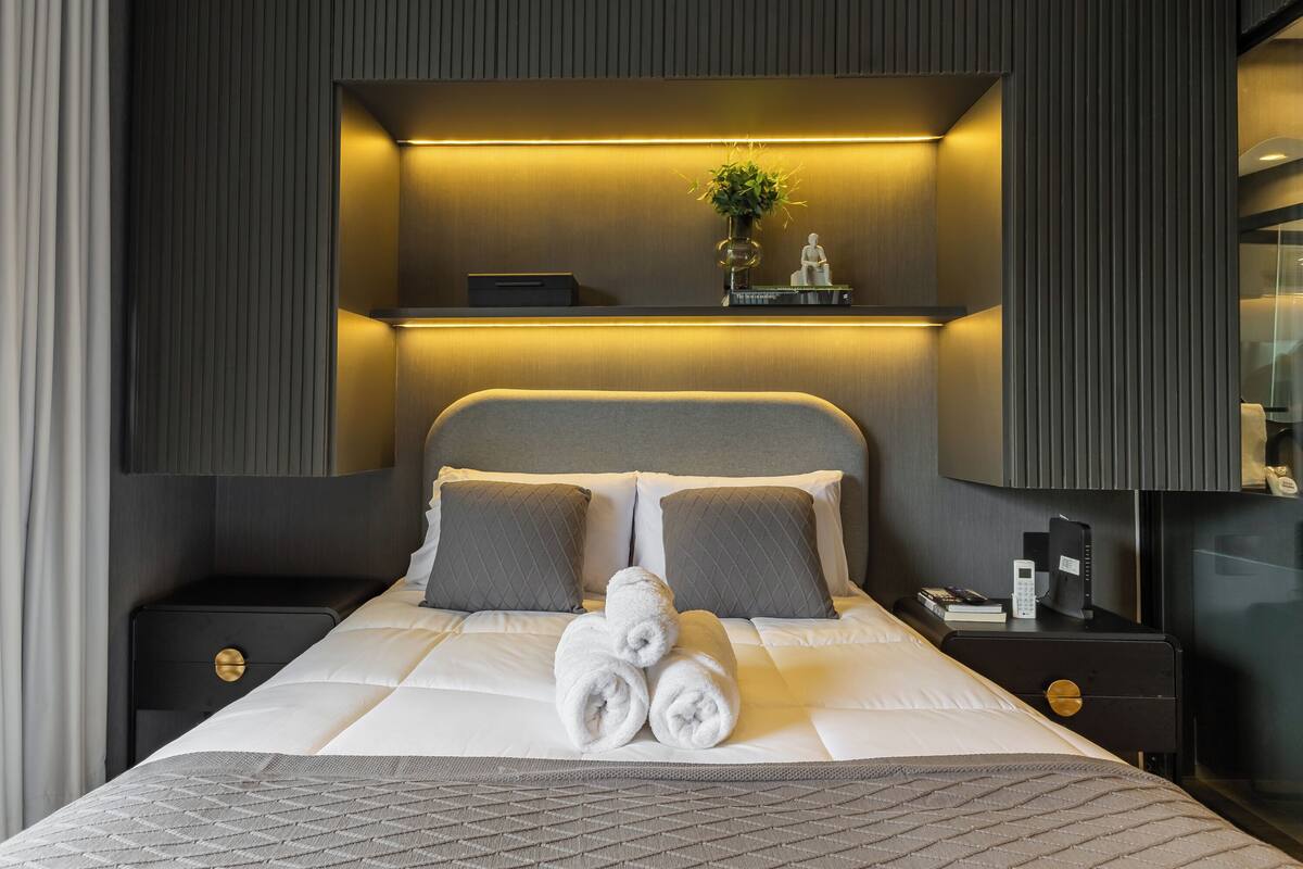 A neatly made bed with pillows, a bedside table with a phone and books, and a wall-mounted shelf with a plant and a statue.