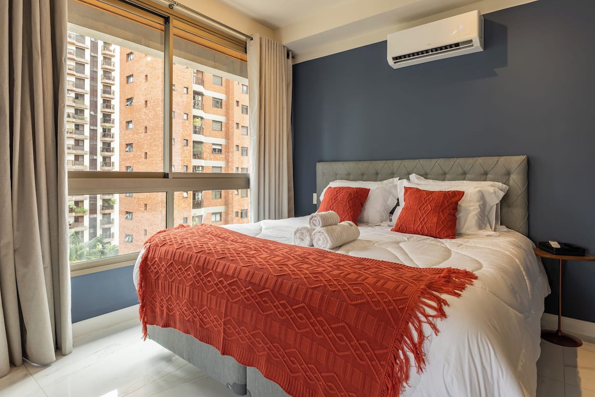 A hotel room with a bed, a window with curtains, an air conditioning unit, and a view of buildings outside.