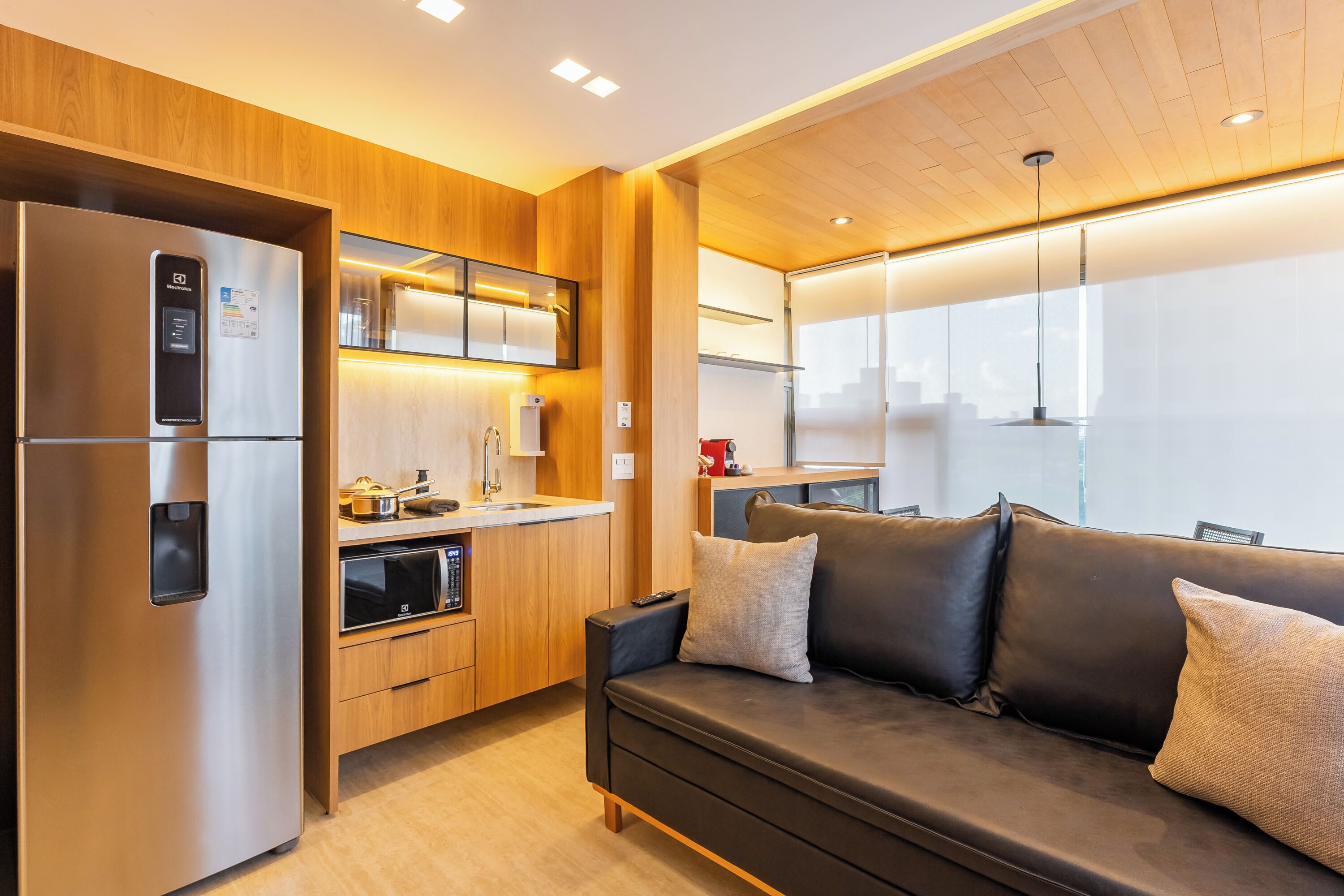 A modern kitchen with a built-in refrigerator, oven, and microwave. There is a wooden panel wall, a sofa with cushions, and a window with a view of the city.