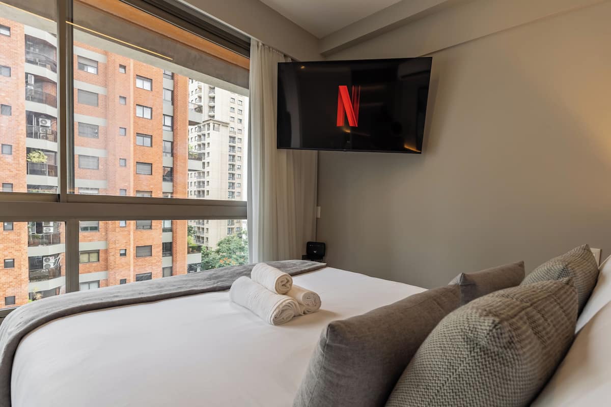 A hotel room with a large bed, a flat-screen TV mounted on the wall, and a view of a cityscape through the window.
