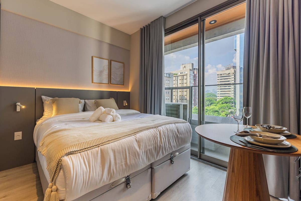 Une chambre d’hôtel moderne avec un grand lit, une table à manger ronde et une vue sur la ville par la fenêtre.