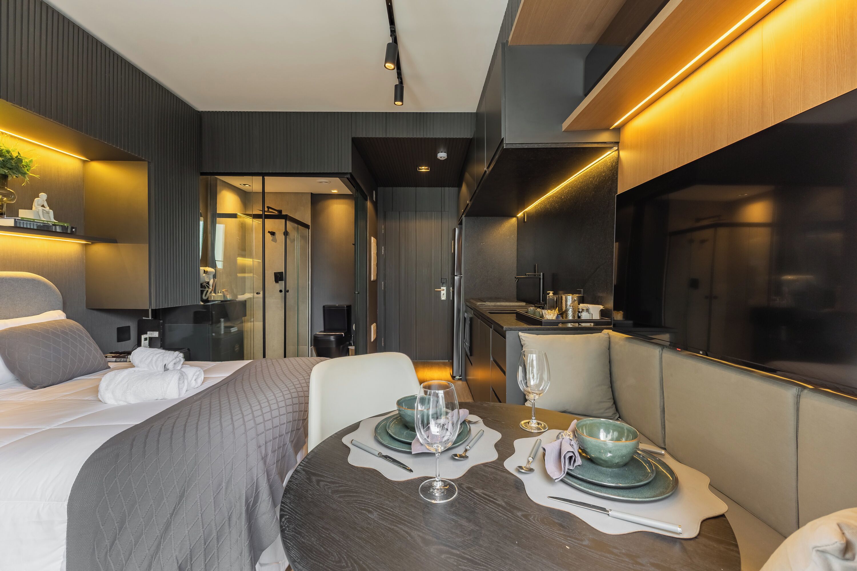 Una habitación de hotel moderna con una cama bien hecha, una mesa de comedor lista para dos personas y una cocina visible a través de una puerta abierta.