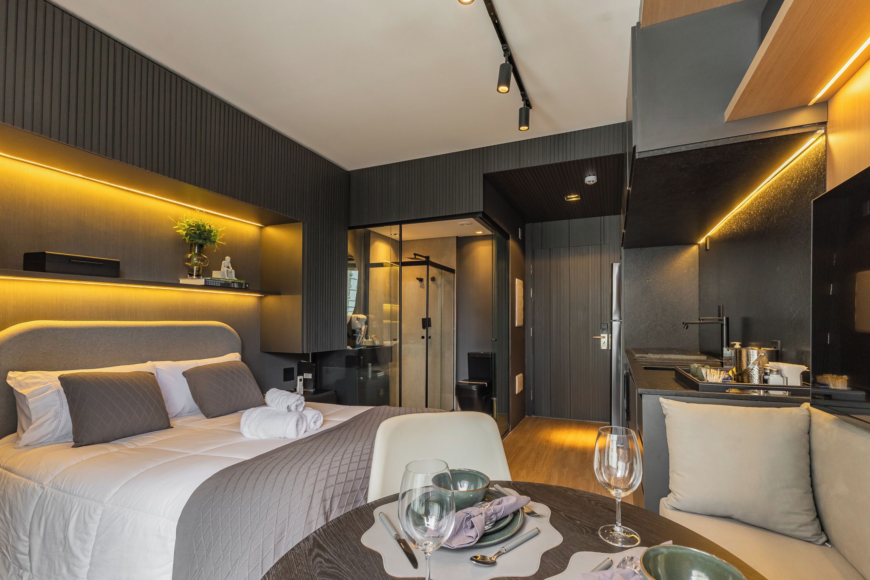 A modern hotel room with a bed, bedside table, and a dining area with a table set for two.