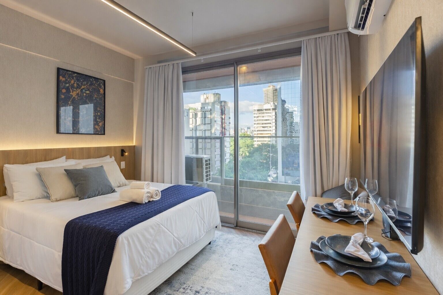 Habitación de hotel con una cama grande, una mesa de comedor con vasos y cubiertos, un televisor de pantalla plana y vistas a la ciudad a través de amplios ventanales.