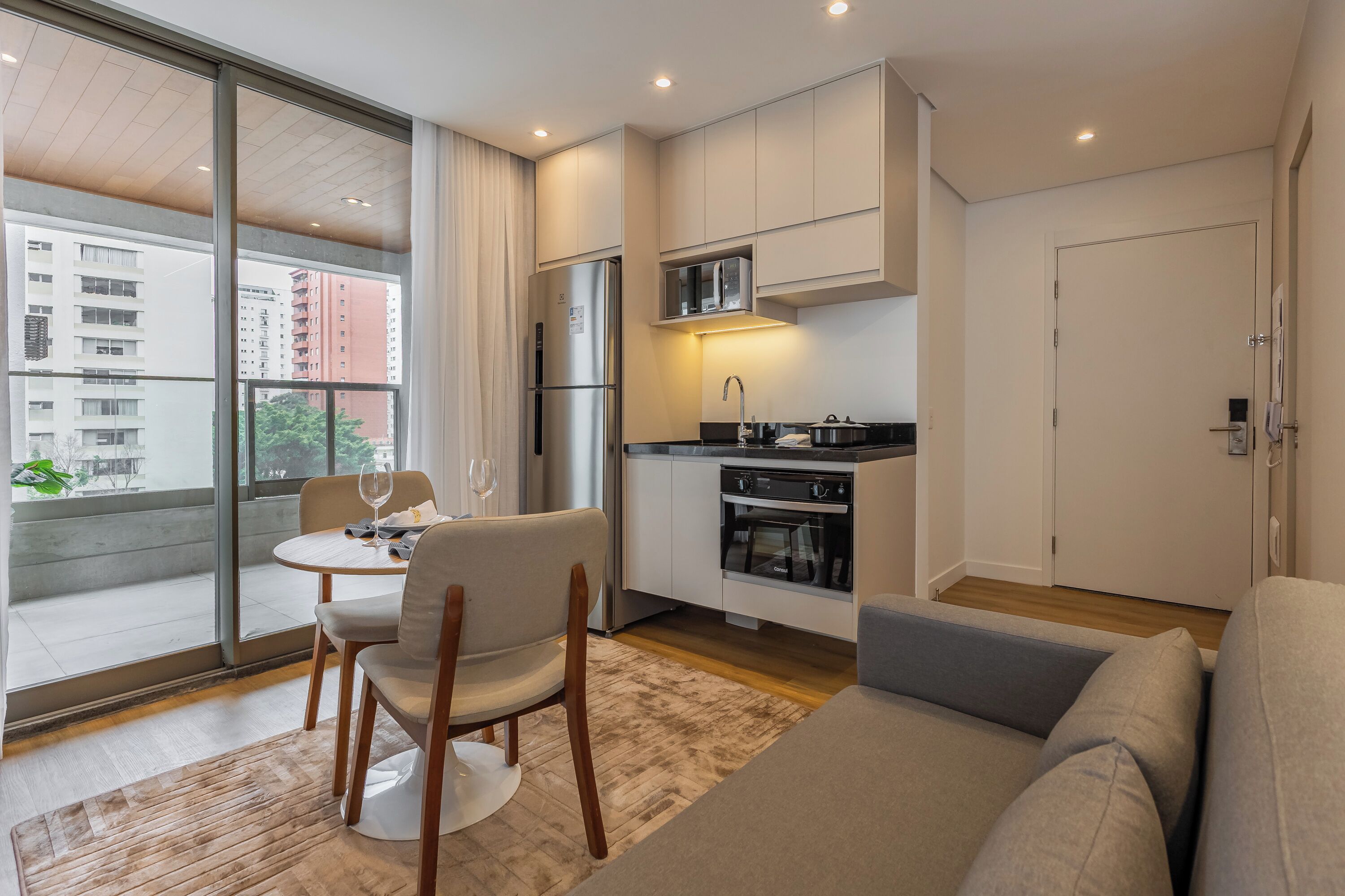 A modern apartment with a dining area, kitchenette, and a large window offering a view of a building.