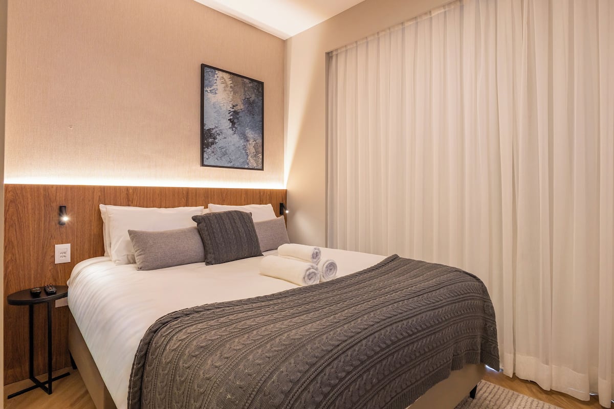 A neatly made bed with a patterned blanket, a headboard with wall-mounted lights, and a large window with blinds.