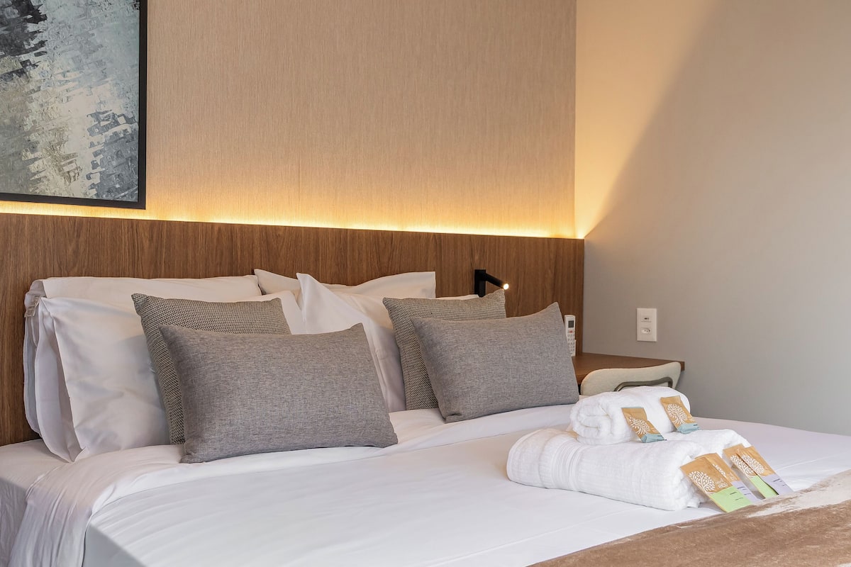 Un lit bien fait, avec du linge de lit blanc et gris, une tête de lit en bois clair et un luminaire fixé au mur.