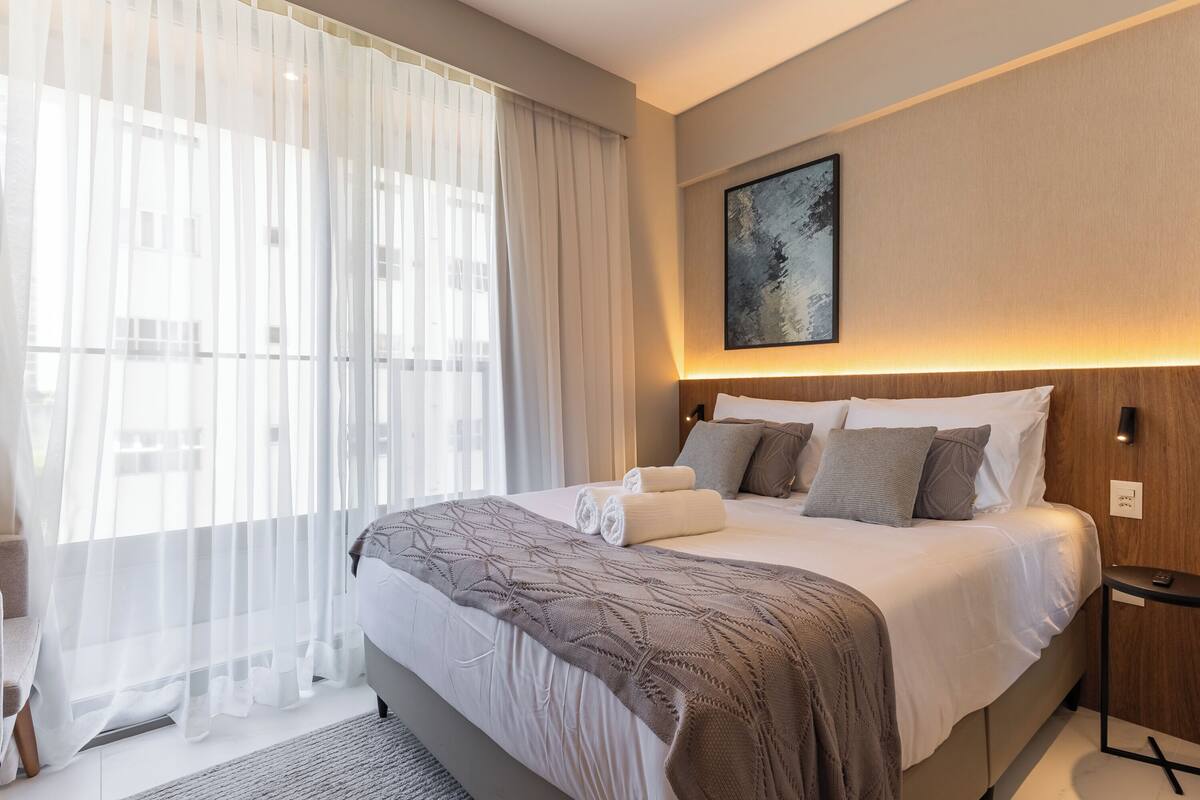 A hotel room with a large bed, a bedside table, a wall-mounted artwork, and sheer curtains.
