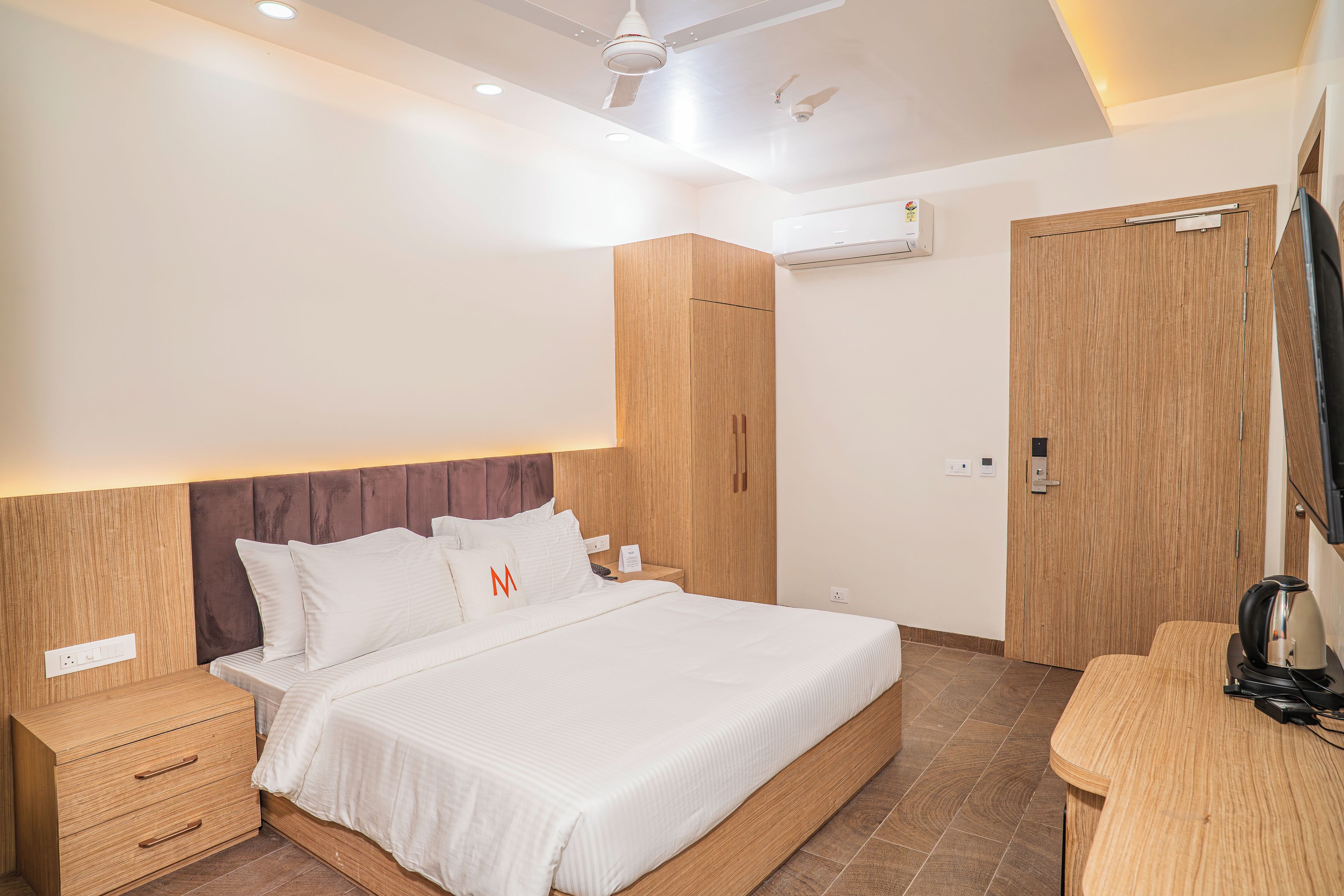 A hotel room with a bed, wooden wardrobe, a bedside table, an air conditioning unit, and a television mounted on the wall.